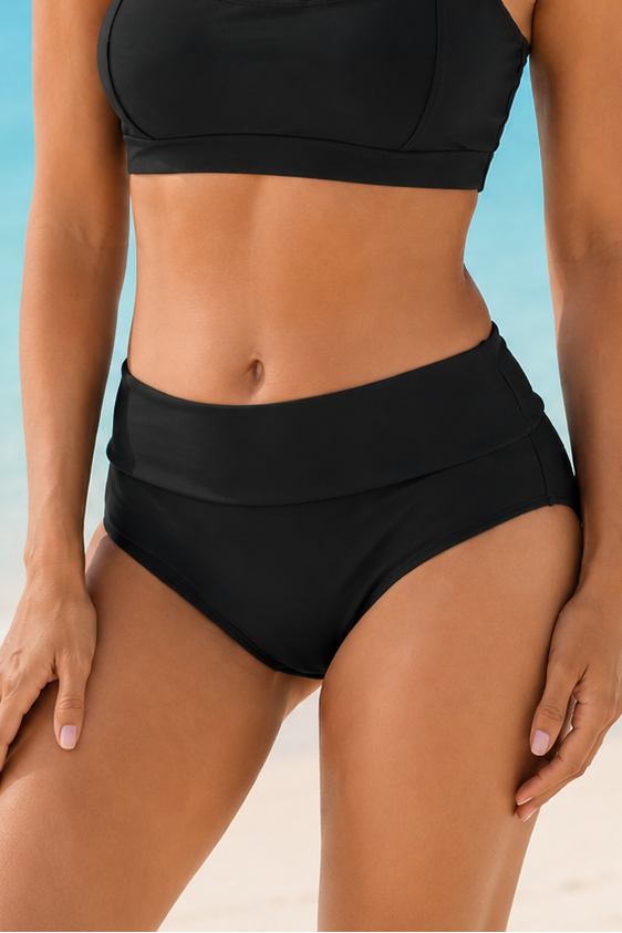 A woman modeling a black two-piece bikini on a sunny beach with turquoise ocean water and pale sand in the background.